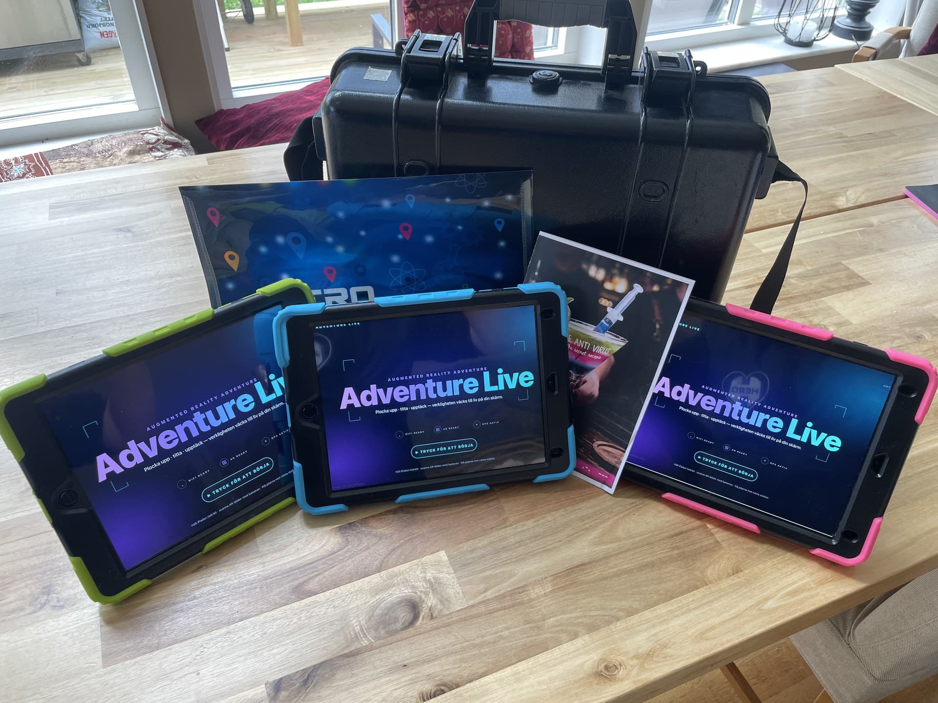 Three iPads in green, blue and pink cases showing the Adventure Live splash, with a black transport case behind them